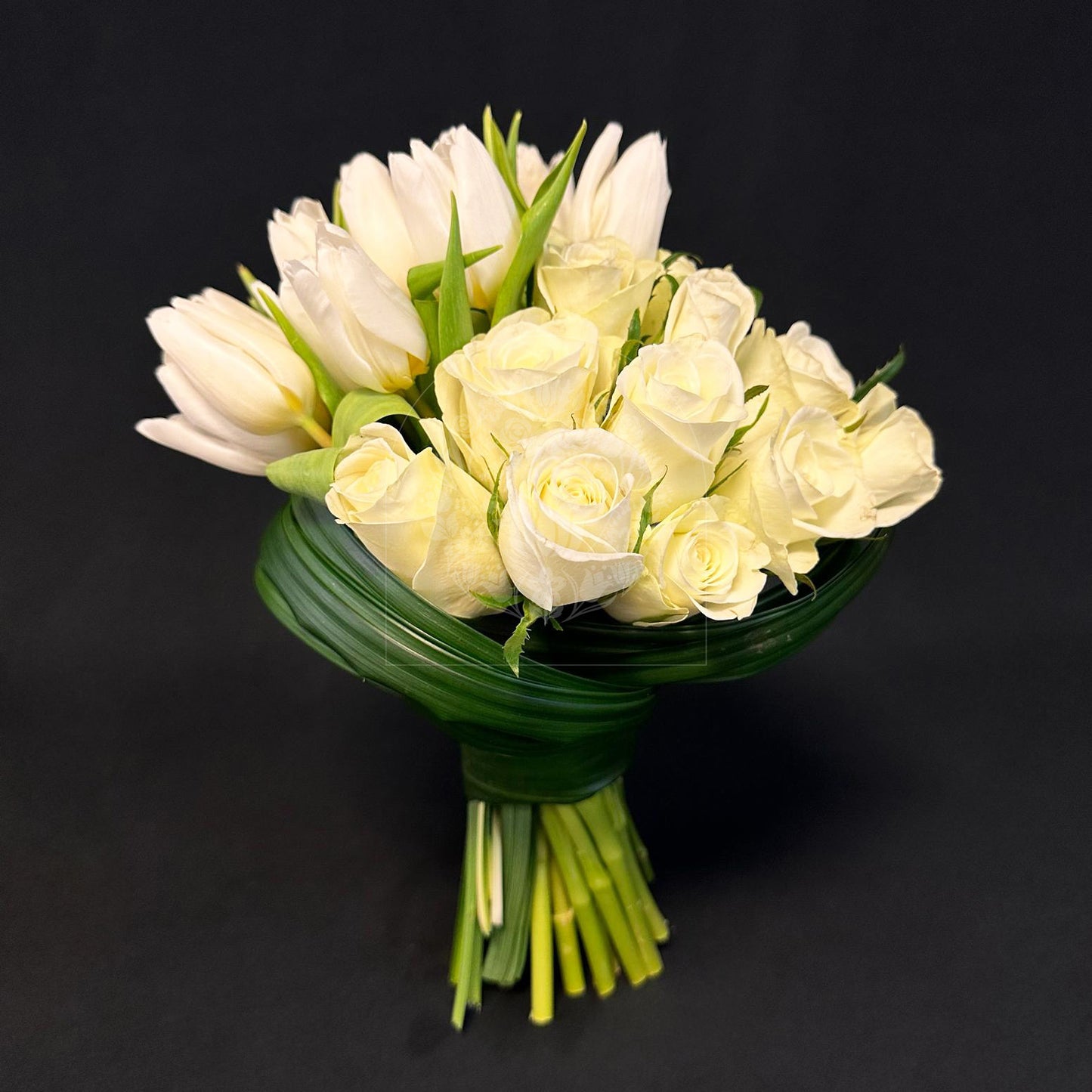 A classic bridal bouquet featuring pristine white roses and tulips, tightly arranged with a wrap of green leaves.