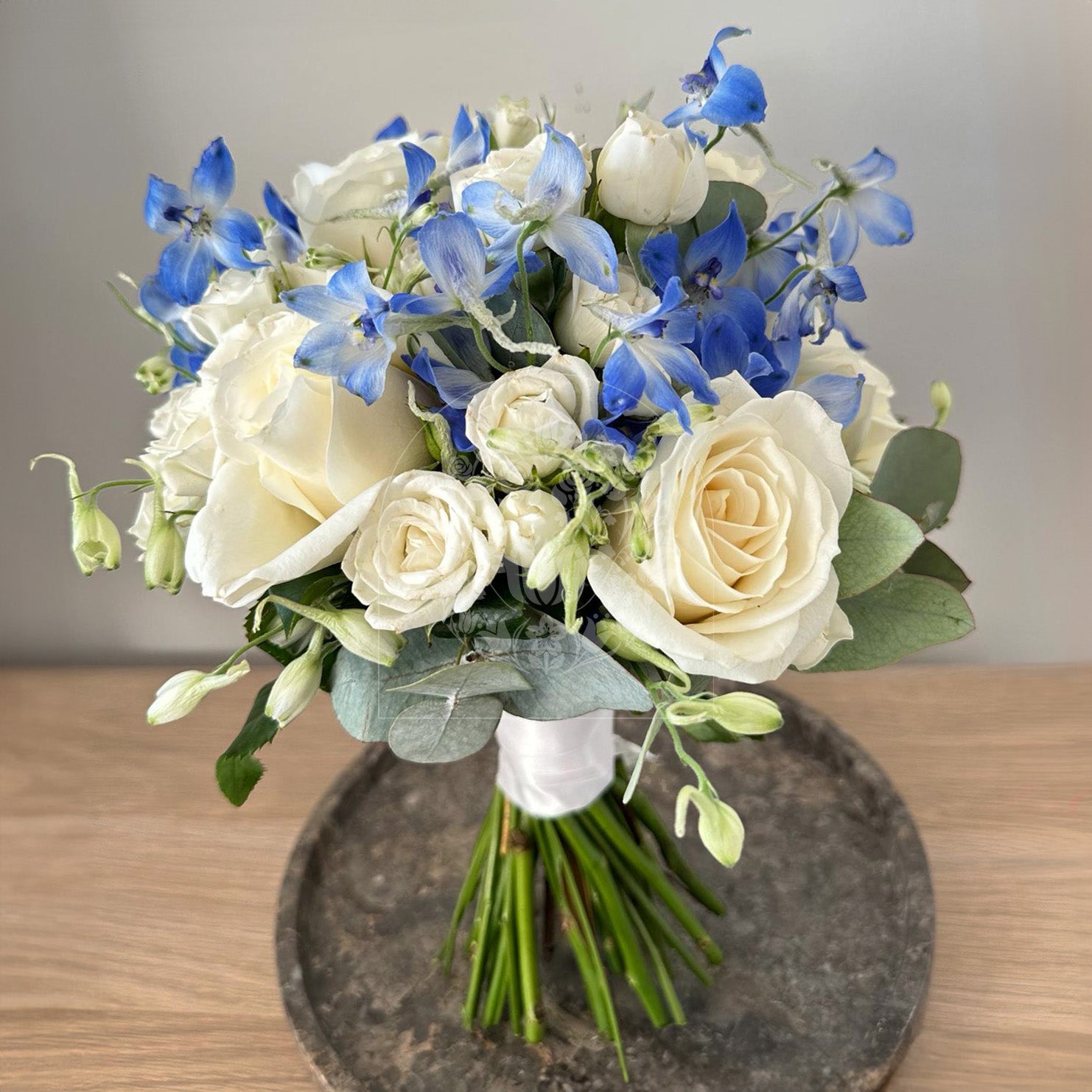 A serene bridal bouquet combining cream roses with delicate blue delphiniums and green foliage, wrapped in a chic white ribbon.